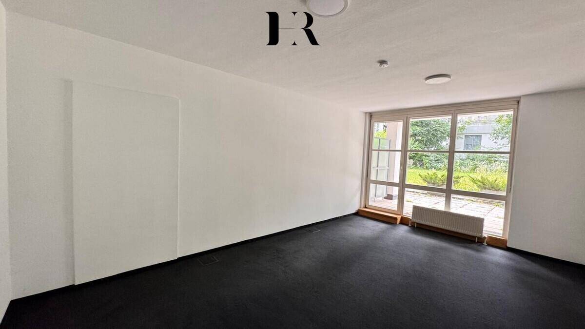 Unmöblierter Raum mit dunklem Teppichboden, weißen Wänden und großen Fenstern zum Außenbereich.