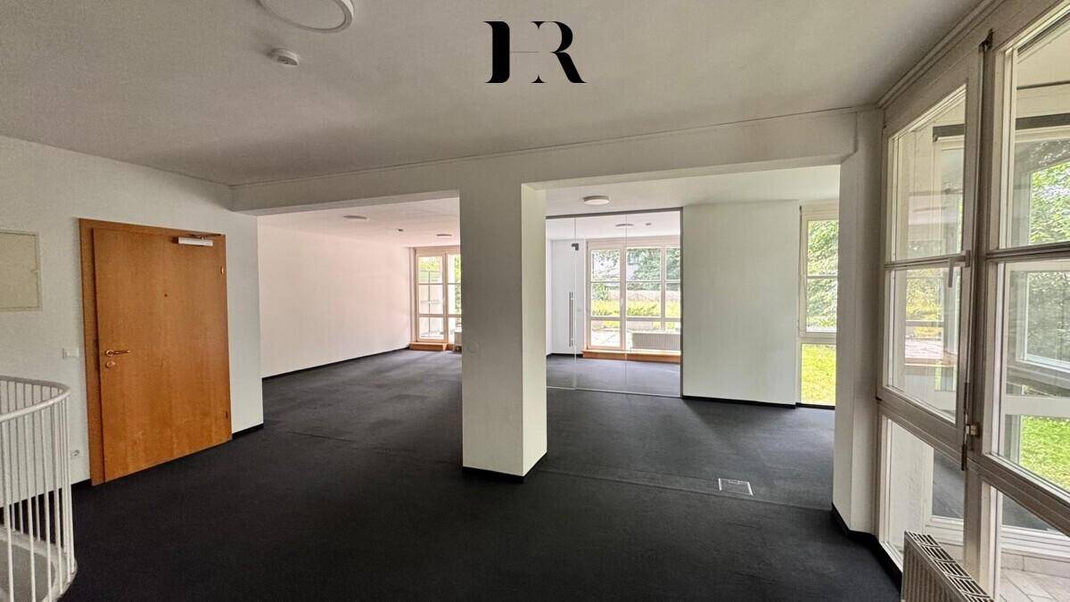 Großer, unmöblierter Raum mit dunklem Teppichboden, großen Fenstern und Wendeltreppe.