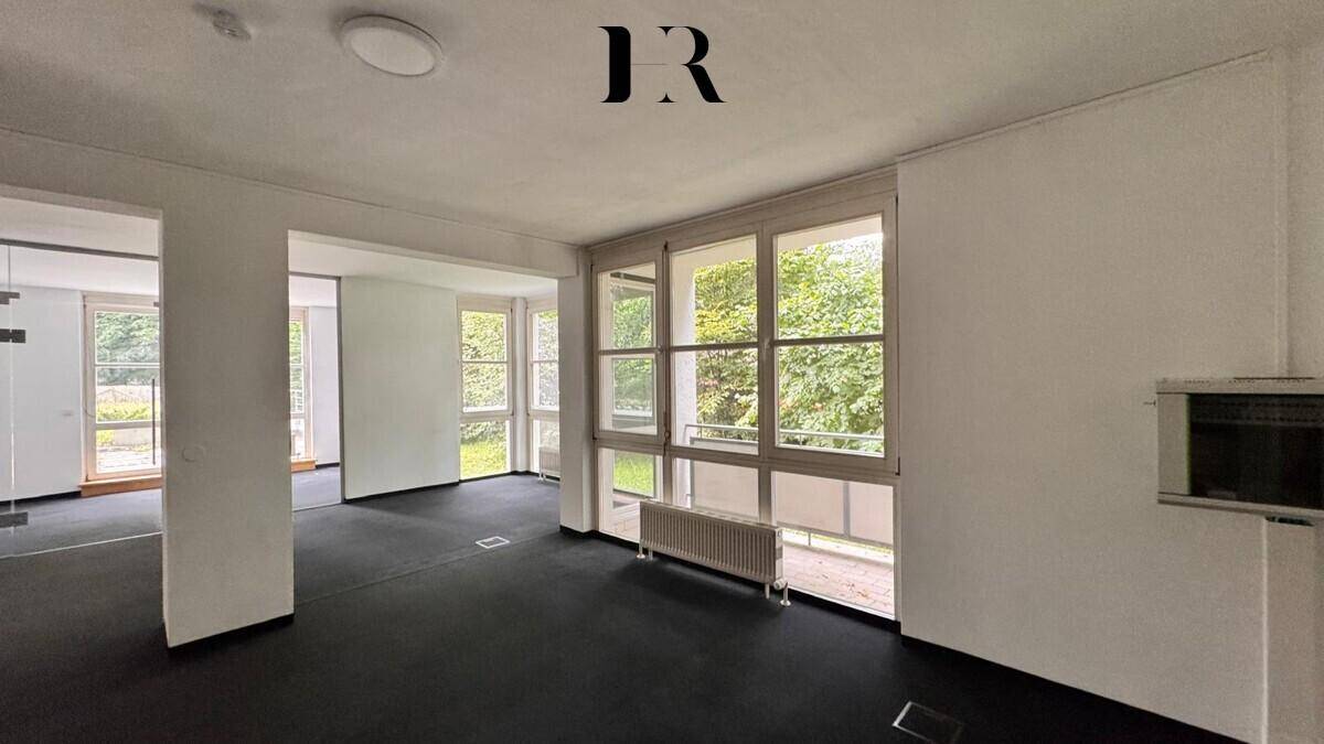 Geräumiger, unmöblierter Raum mit dunklem Teppichboden, großen Fenstern und Blick ins Grüne.