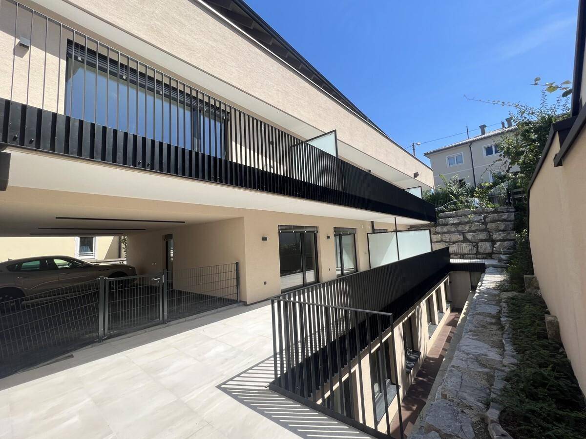 Moderne Gebäudefassade mit Balkonen, Carport-Einfahrt und einer unteren Terrassenebene.