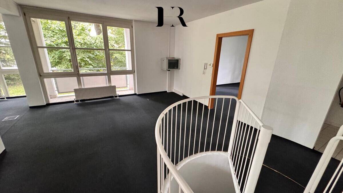 Geräumiger, unmöblierter Raum mit dunklem Teppichboden, großen Fenstern und Wendeltreppe.