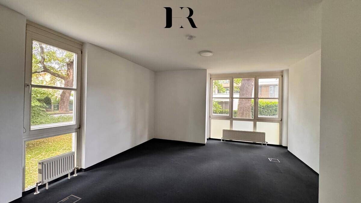 Heller, unmöblierter Raum mit dunklem Teppichboden, weißen Wänden und zwei großen Fenstern.