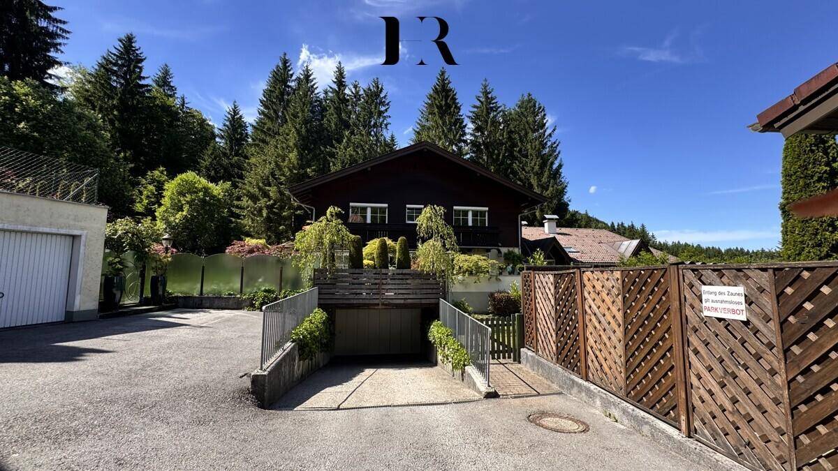 Dunkles Holzhaus mit integrierter Garage und Auffahrt, umgeben von Bäumen und einem Holzzaun.