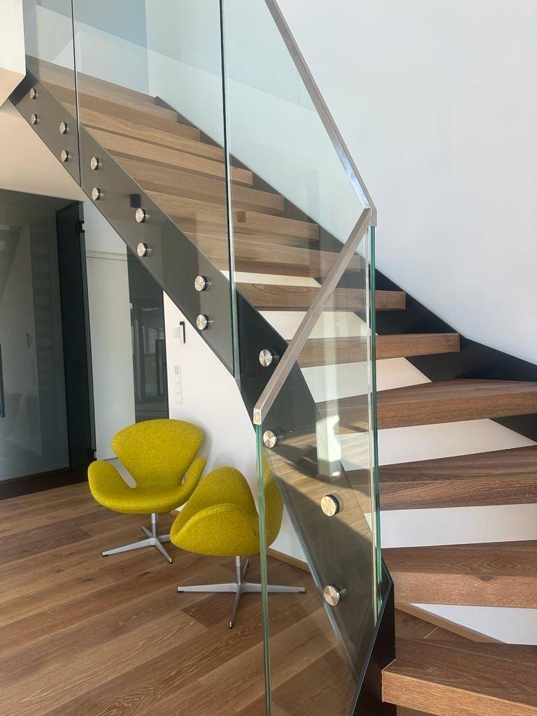 Elegante Holztreppe mit Glasgeländer und zwei modernen gelben Sesseln im offenen Wohnbereich.