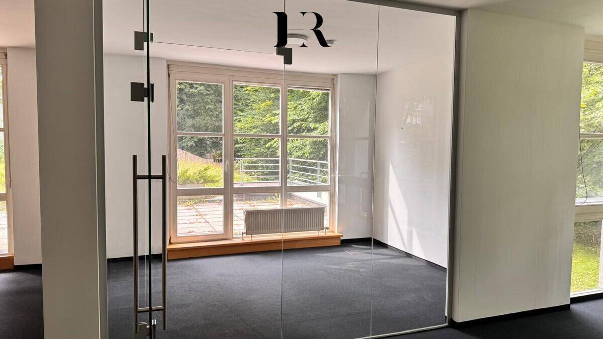 Heller Büroraum mit Glaswänden, dunklem Teppichboden, großen Fenstern und Blick ins Grüne.
