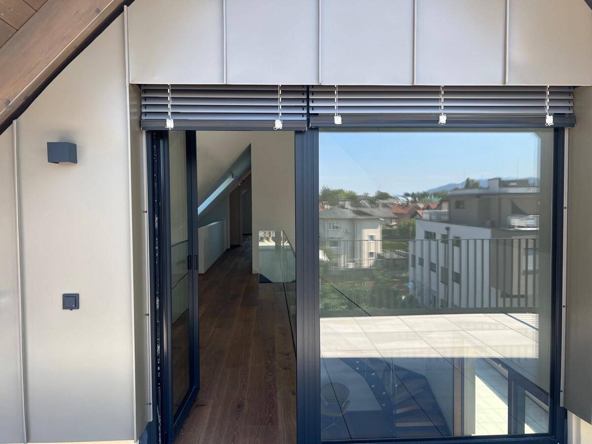 Blick aus einem Innenraum durch große Fenster auf eine sonnige Terrasse und die Umgebung.