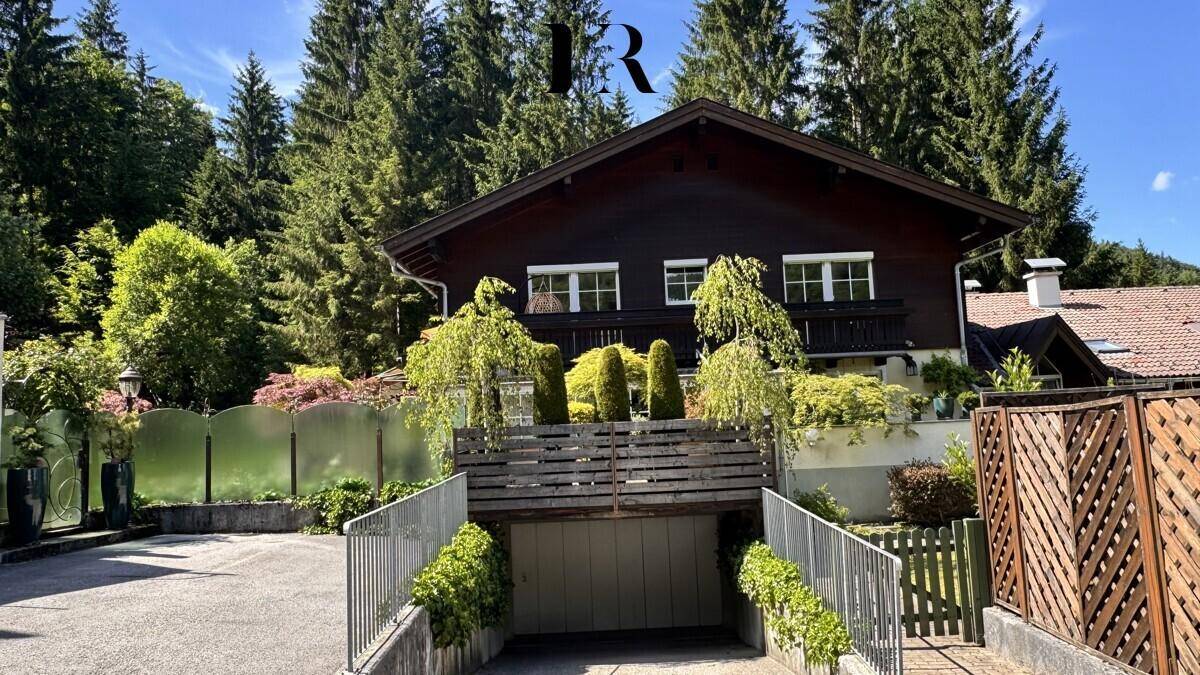 Charmantes Holzhaus mit Satteldach und Tiefgaragenzufahrt, eingebettet in eine grüne Umgebung.