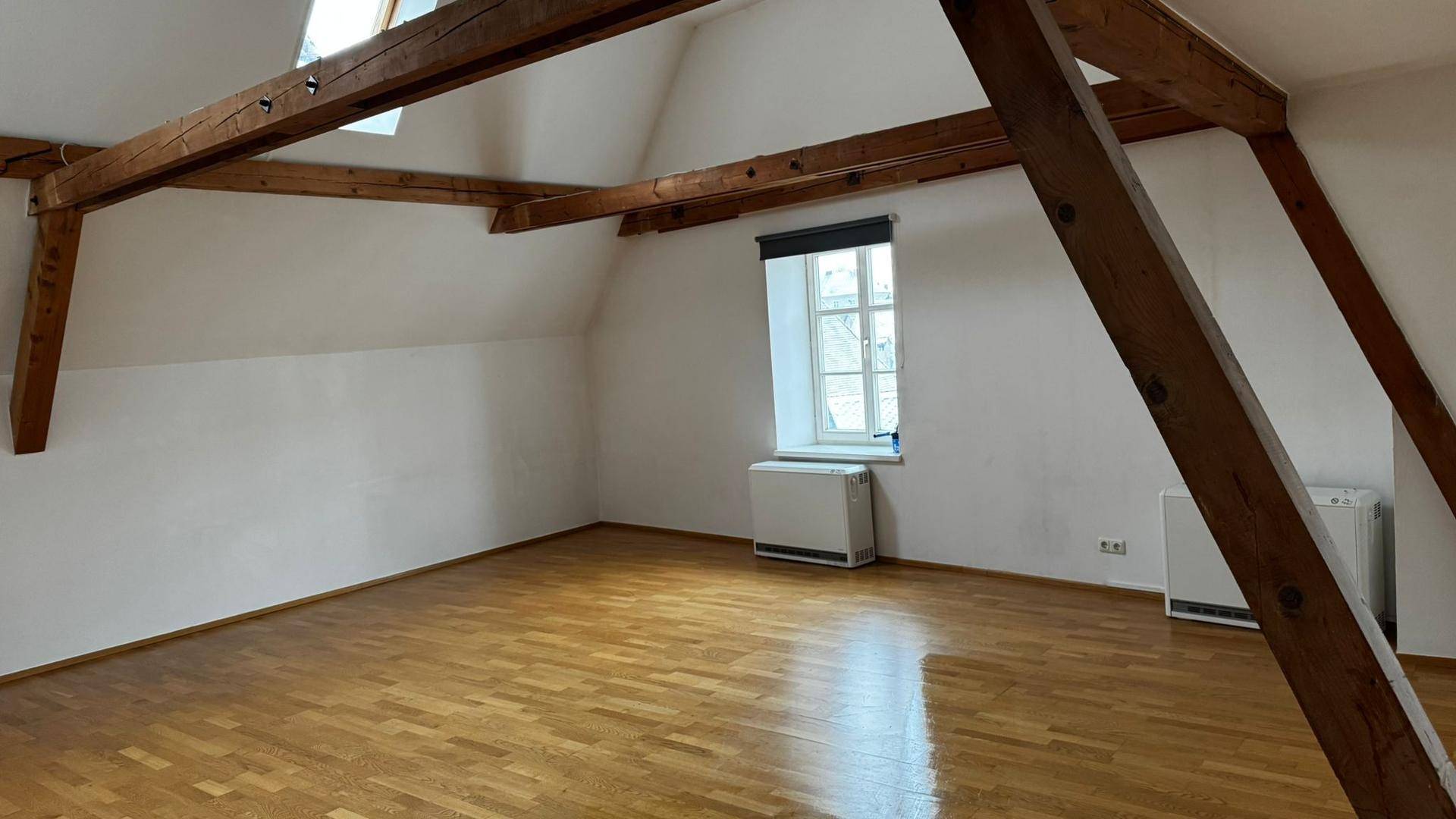 Geräumiger Dachgeschossraum mit Parkettboden, sichtbaren Holzbalken, Dachfenstern und einem traditionellen Fenster.