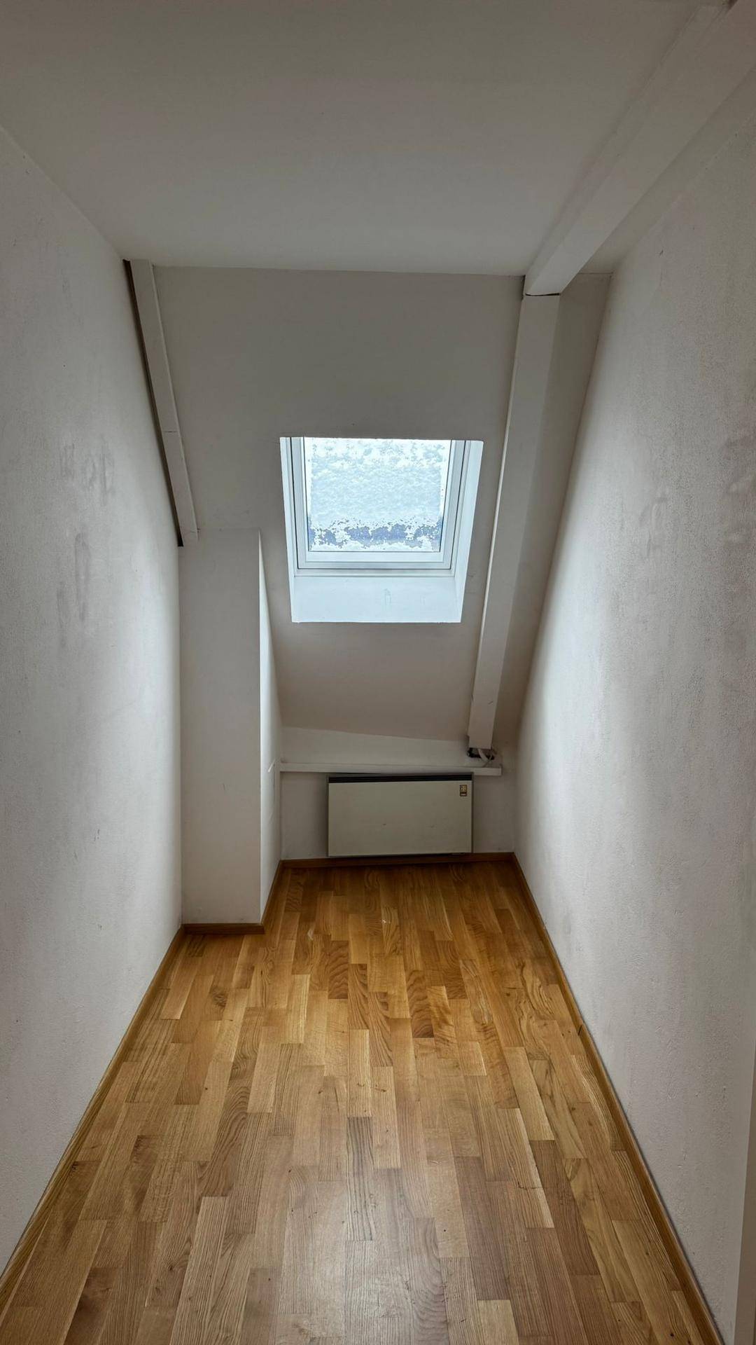 Schmaler Raum im Dachgeschoss mit Parkettboden, Dachfenster und Heizkörper unter der Schräge.