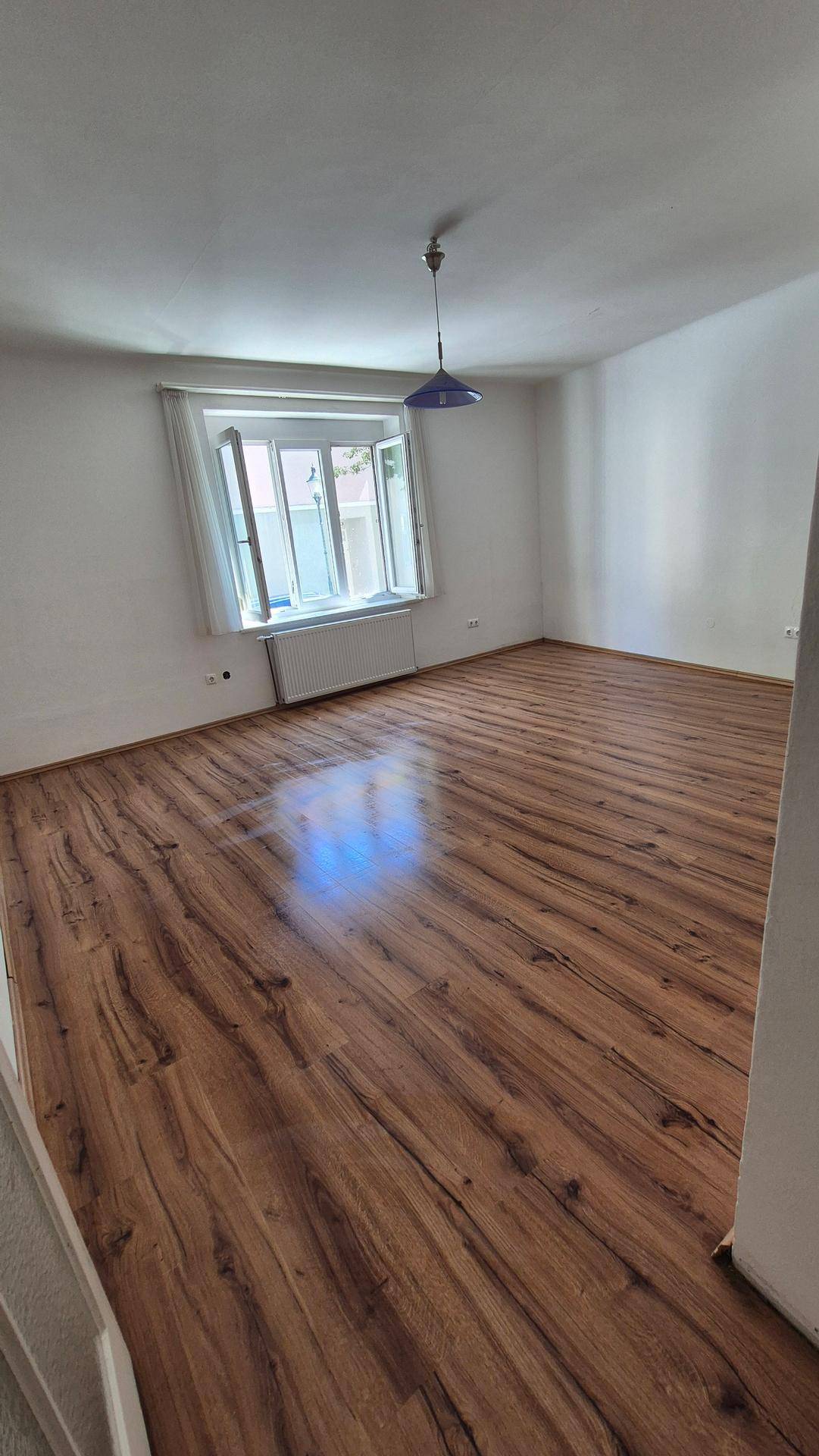 Helles Zimmer mit Laminatboden, weißen Wänden und einer auffälligen blauen Pendelleuchte.
