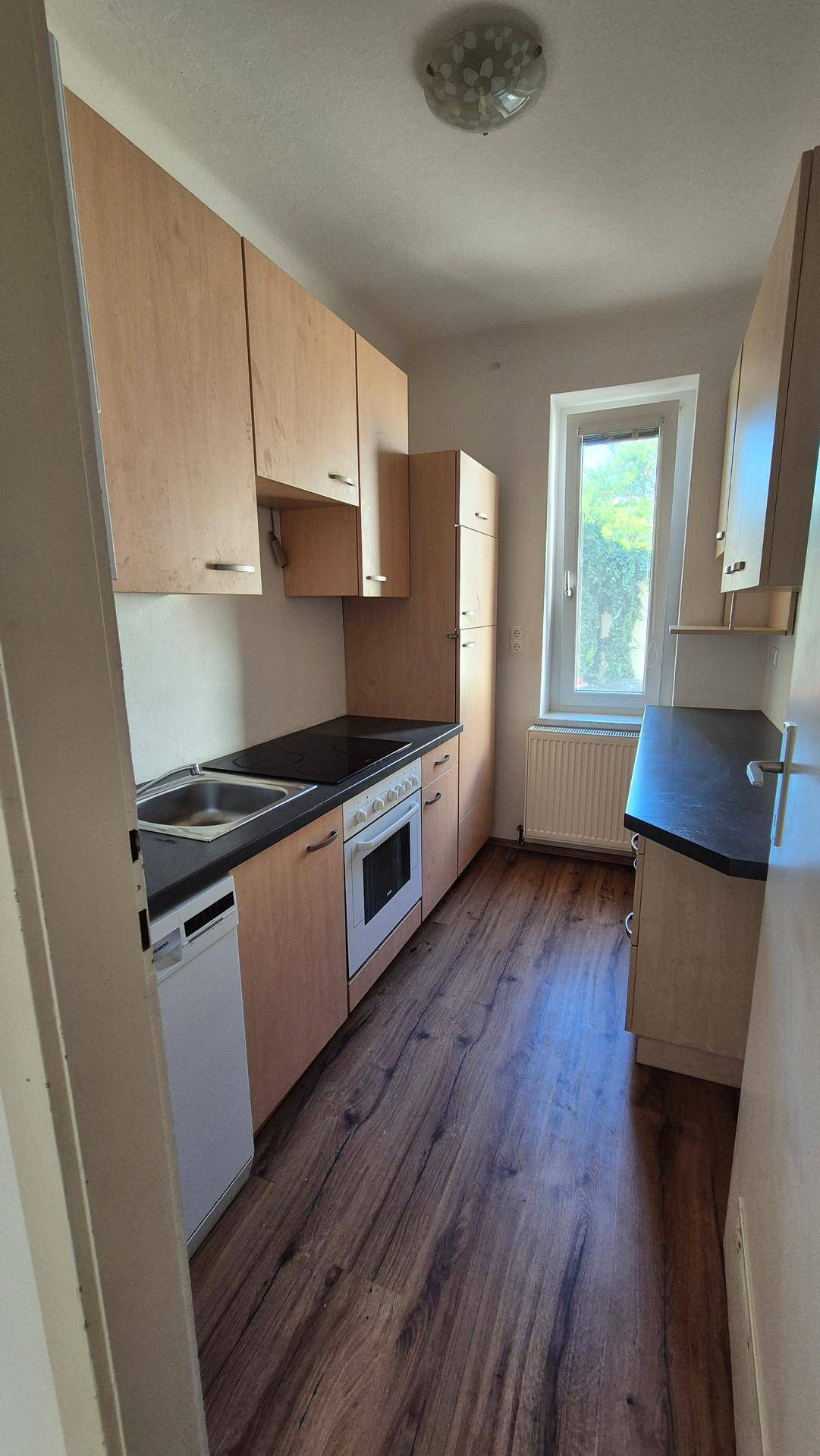Schmale Küche mit Holzschränken, Einbaugeräten und Fenster für Tageslicht.