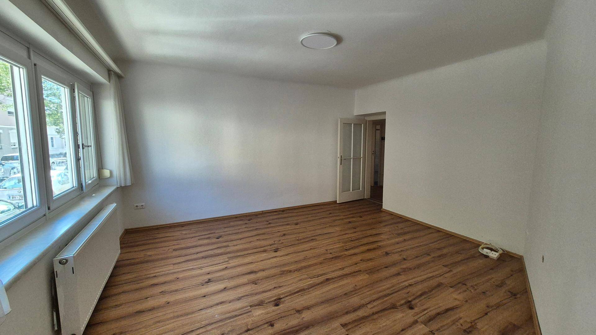 Geräumiges Wohnzimmer mit Laminatboden, weißen Wänden und einer Tür mit Glaseinsätzen.