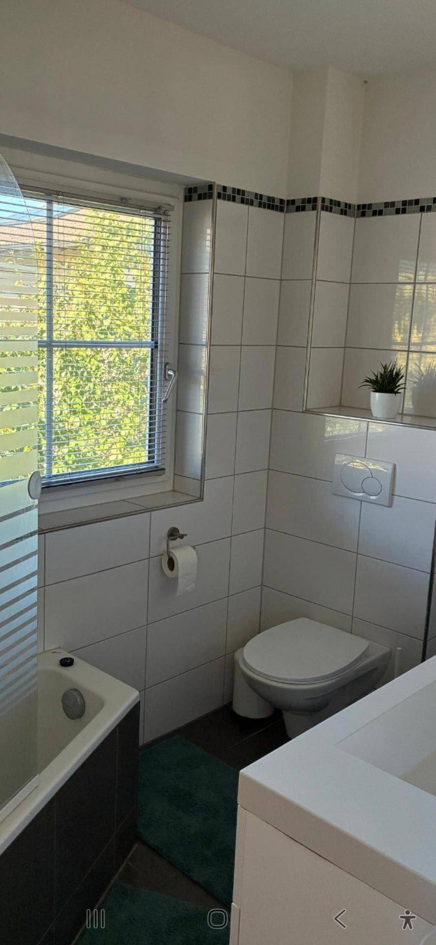 Helles Badezimmer mit weißen Fliesen, Badewanne, WC und Fenster für natürliche Belüftung.