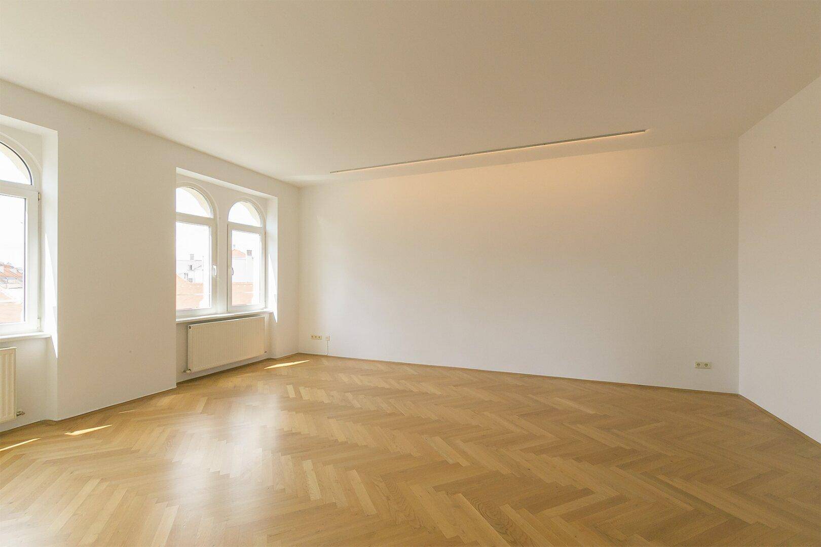 Großzügiges, unmöbliertes Zimmer mit hellem Fischgrätparkett und viel Tageslicht.