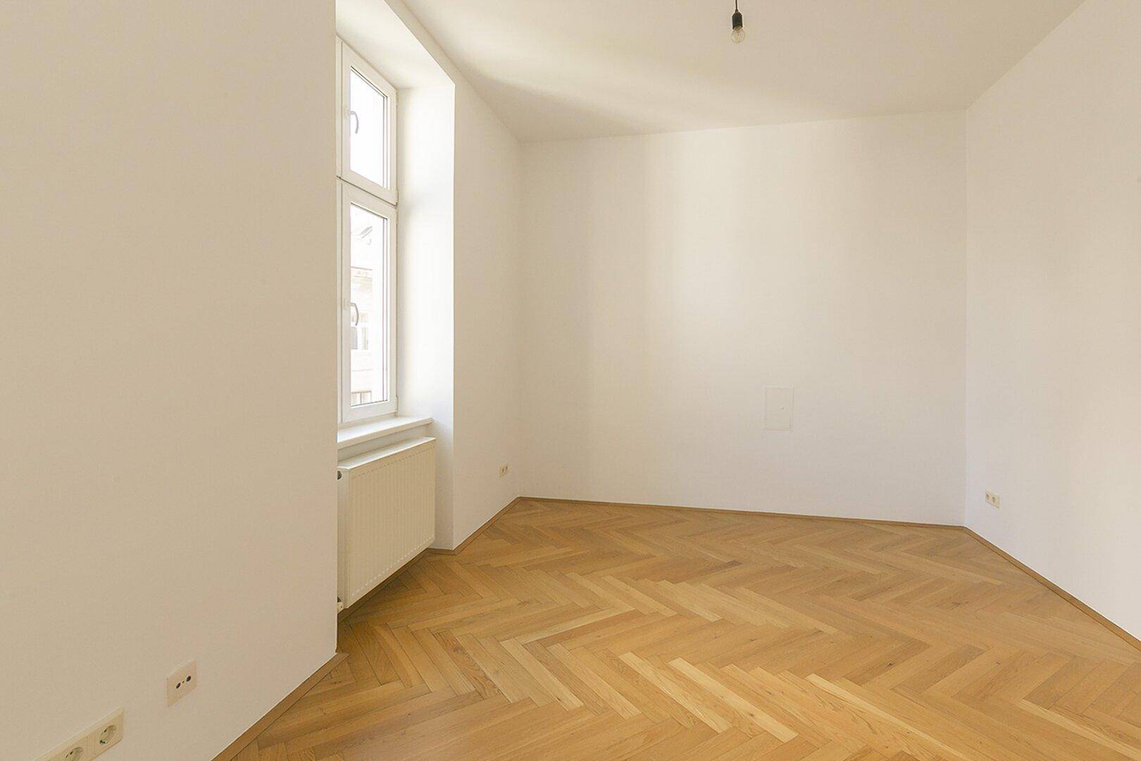 Ruhiges Zimmer mit Fischgrätparkett und einem Fenster, das viel Tageslicht hereinlässt.
