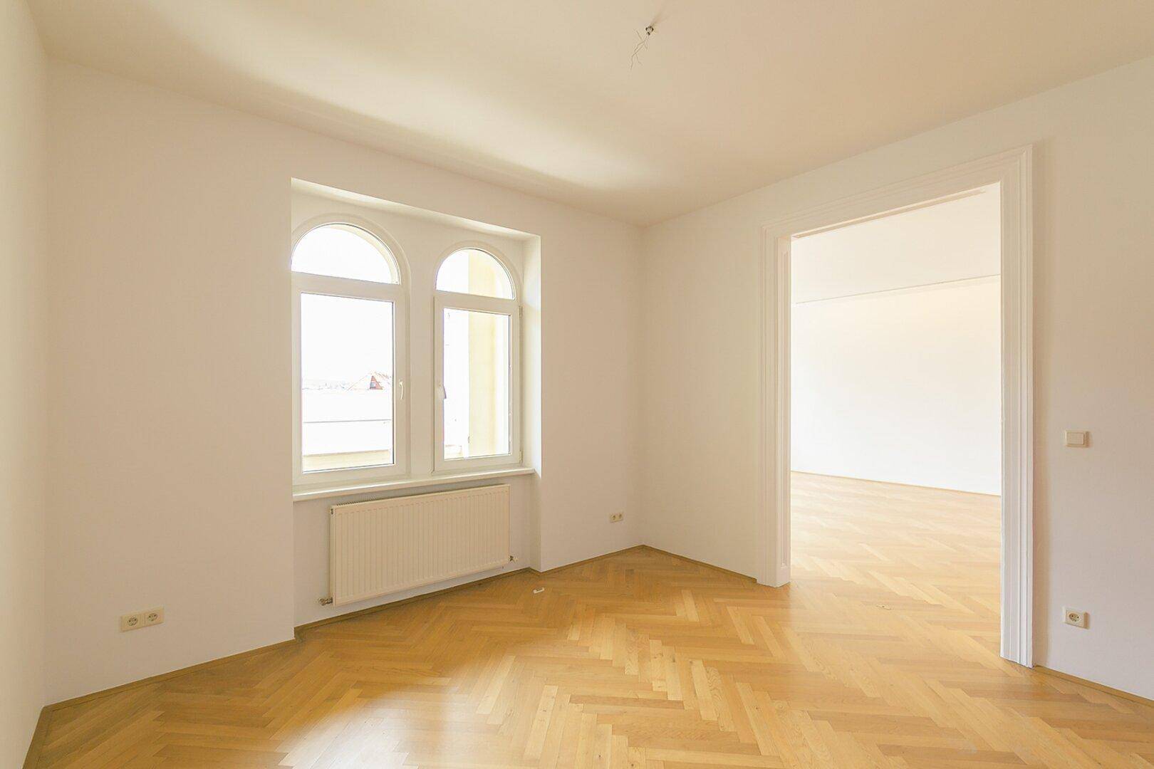 Helles Zimmer mit Fischgrätparkett und Zugang zu einem weiteren geräumigen Wohnbereich.