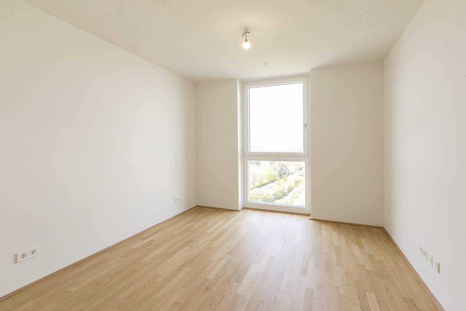 Helles Schlafzimmer mit Parkettboden und einem großen Fenster, das viel Tageslicht in den Raum bringt.