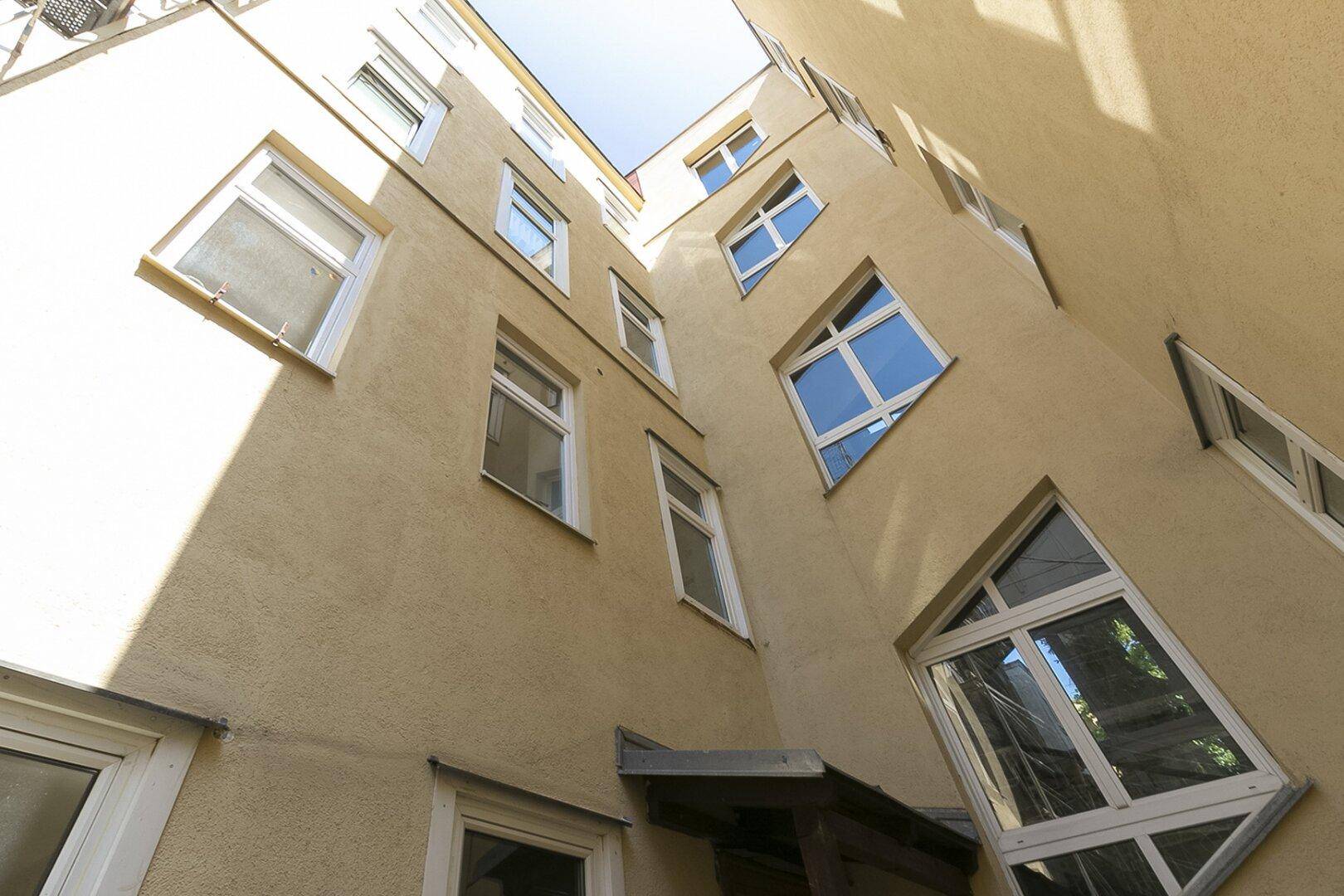 Blick nach oben auf die helle Fassade des Innenhofs mit vielen Fenstern und Sonnenschein.