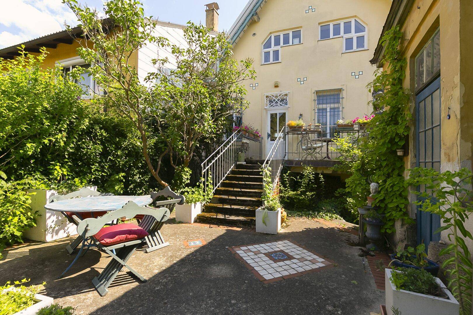 Garten mit gepflastertem Sitzbereich, Treppe zum Haupthaus und üppiger Vegetation.