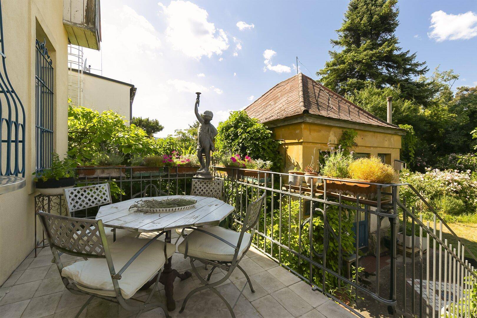 Gemütliche Terrasse mit Gartenmöbeln, Statue und üppiger Bepflanzung im Hintergrund.