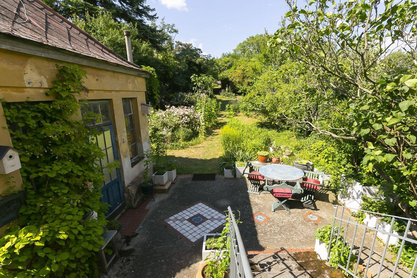 Gepflasterter Terrassenbereich im Garten mit Sitzgelegenheiten und Blick auf ein Nebengebäude.