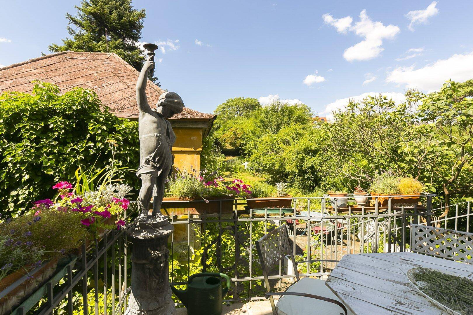 Sonniger Terrassenbereich mit Gartenmöbeln, Statue und weitem Blick auf die grüne Umgebung.
