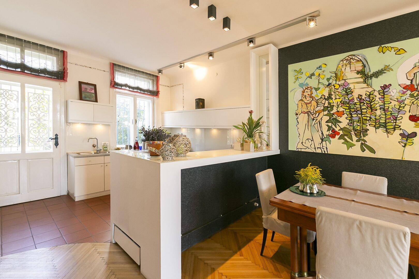 Offene Küche mit Essbereich, weißer Theke und farbenfrohem Wandbild im Hintergrund.