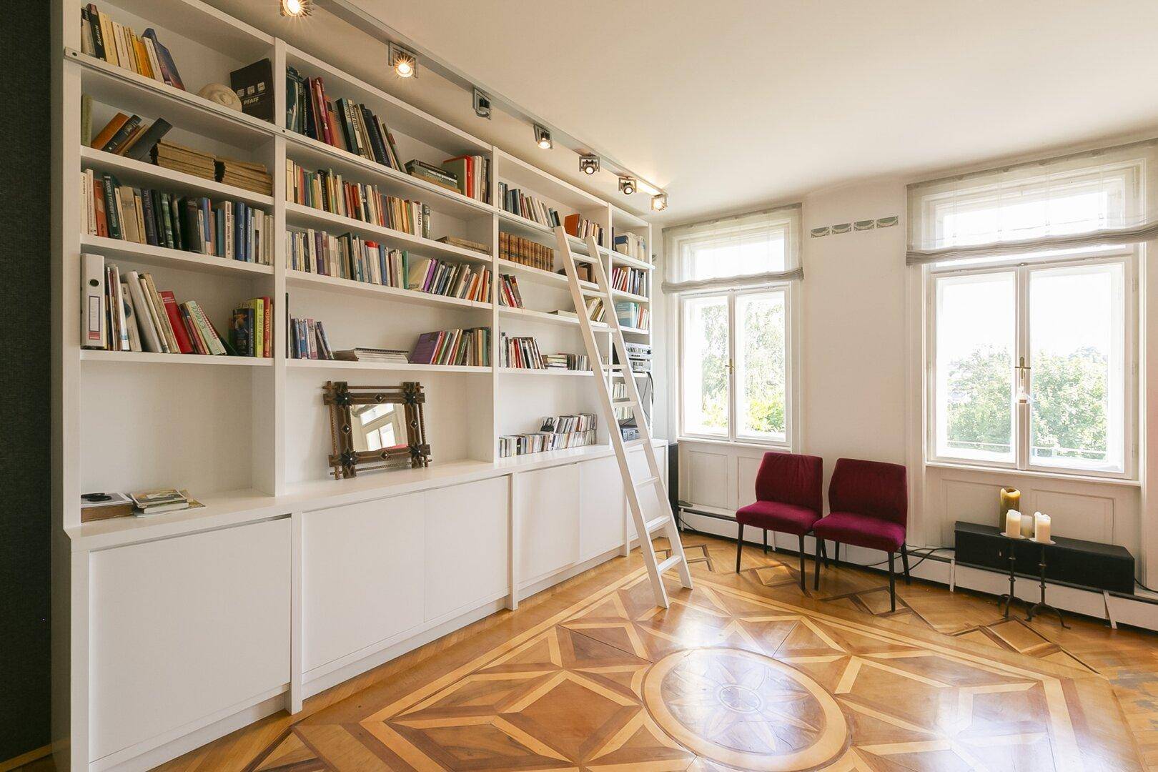 Großes Bücherregal mit Leiter in einem hellen Raum mit gemustertem Parkettboden und Fenstern.