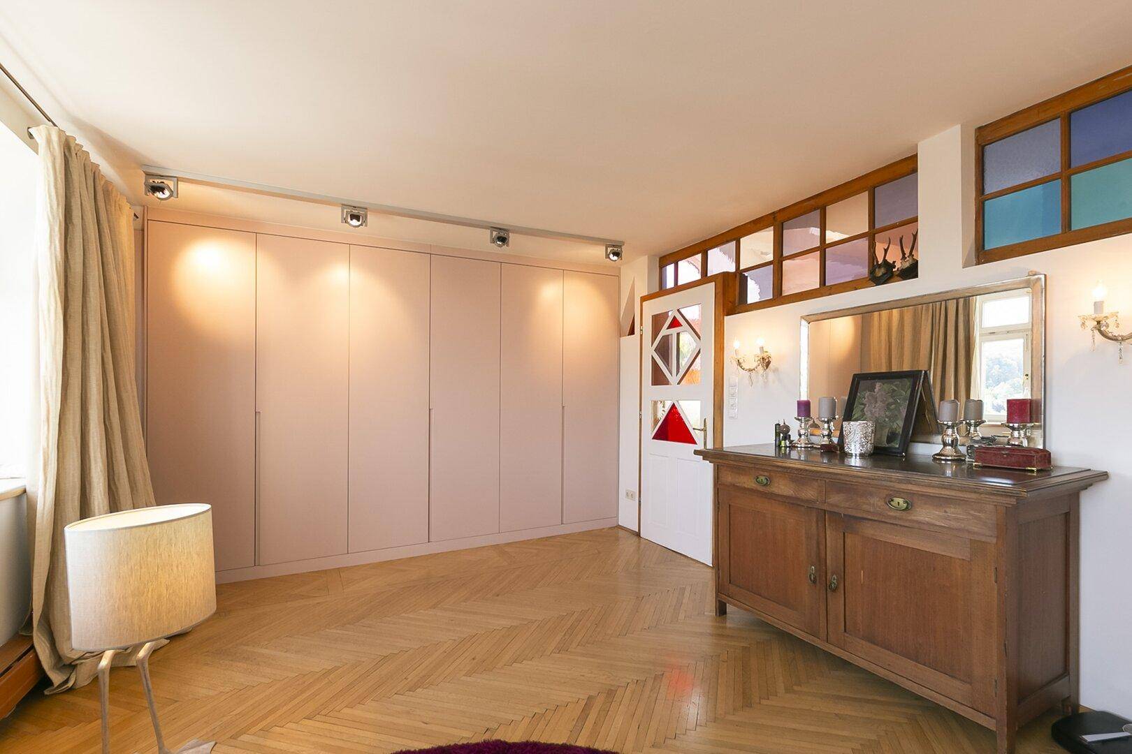 Schlafzimmer mit Einbauschränken, Fischgrätparkett und dekorativen Fenstern.