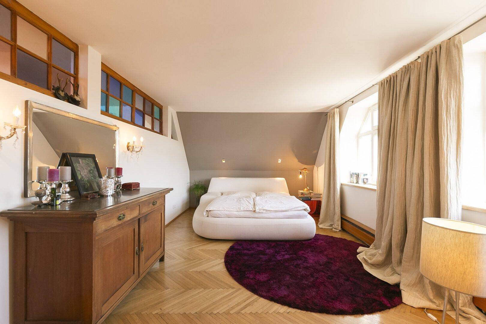 Gemütliches Schlafzimmer mit modernem Bett, Fischgrätparkett und farbigen Fenstern.