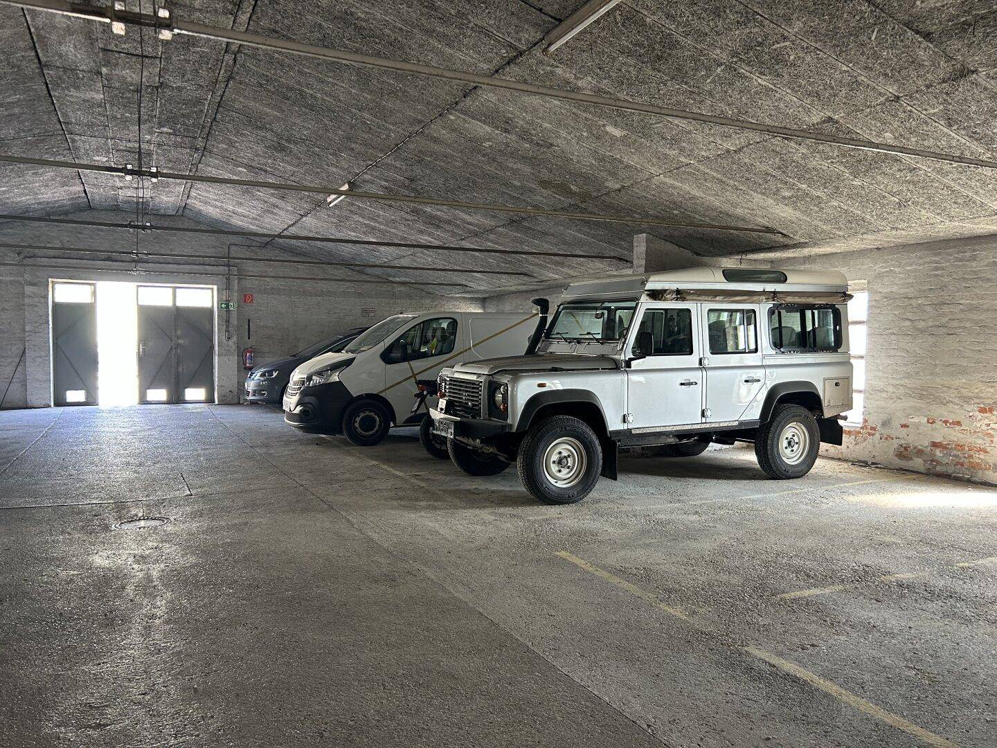 Weitläufige Tiefgarage mit mehreren Fahrzeugen, Betonboden und großen Toren im Hintergrund.