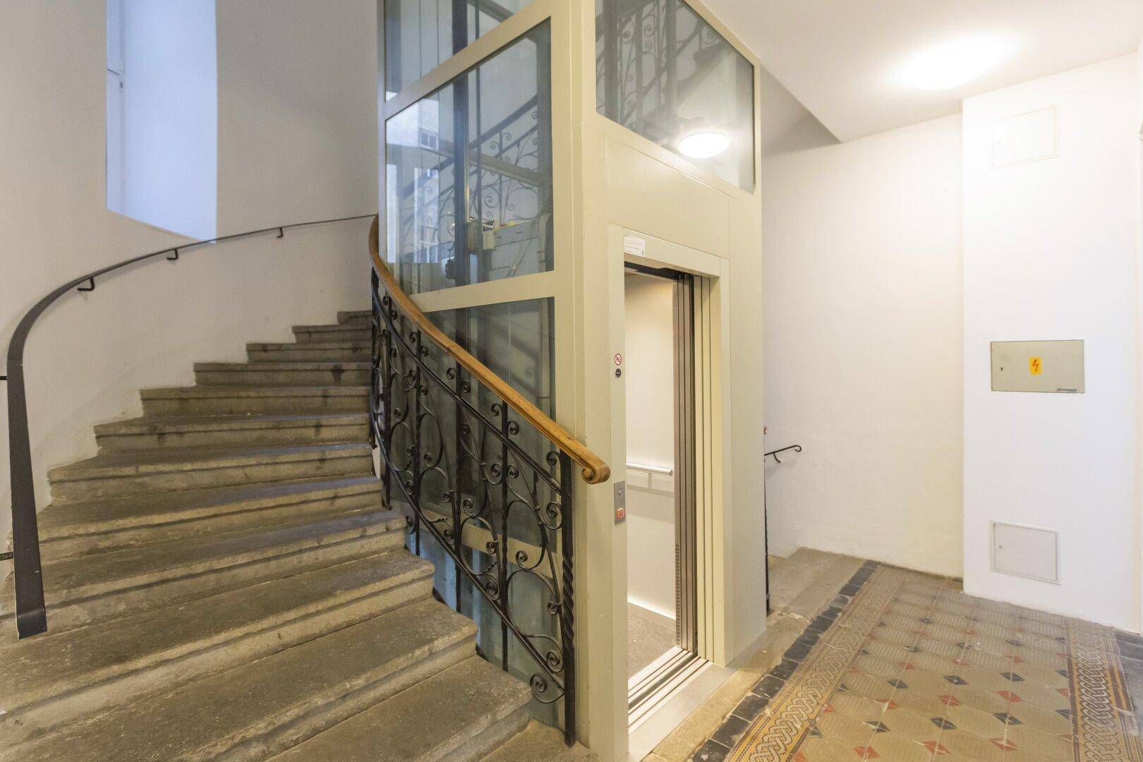 Historisches Treppenhaus mit schmiedeeisernem Geländer und modernem Glasaufzug.