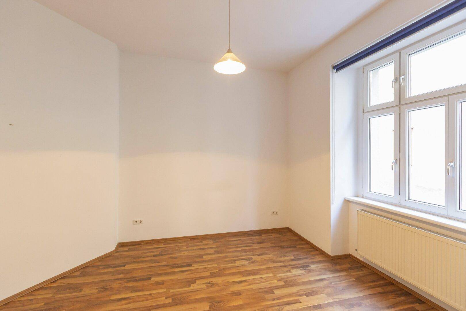 Detailansicht eines hellen Zimmers mit Laminatboden und einem großen Fenster.