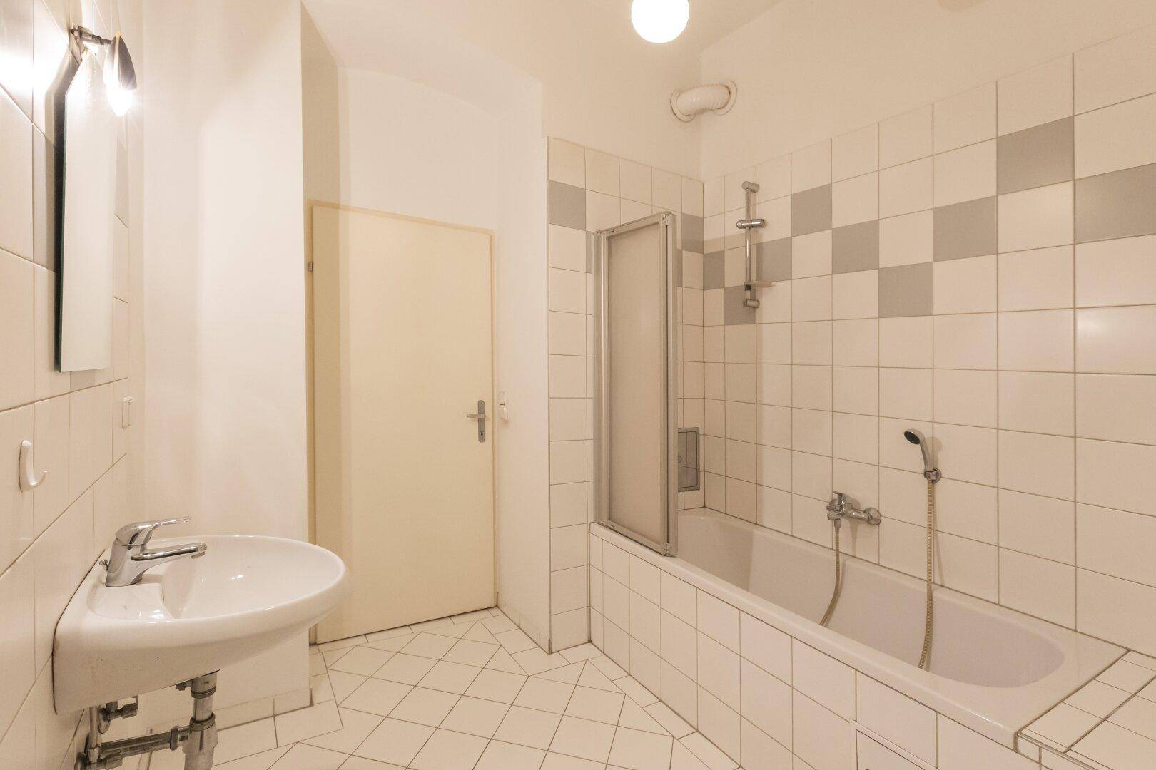 Badezimmer mit Badewanne, Waschbecken und weißen Fliesen, teilweise mit grauen Akzenten.