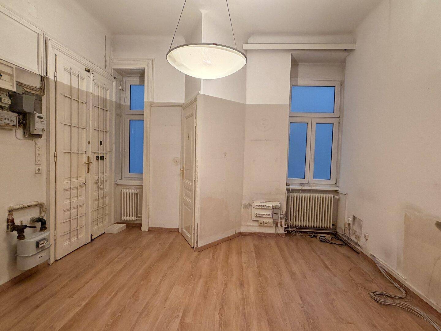 Heller Raum mit Laminatboden und hohen Fenstern, der Renovierungsbedarf aufweist und Versorgungszähler zeigt.
