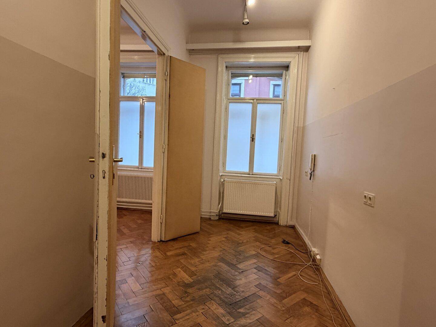 Langer, schmaler Raum mit Fischgrätparkett und zwei Fenstern, der Renovierungsbedarf zeigt.