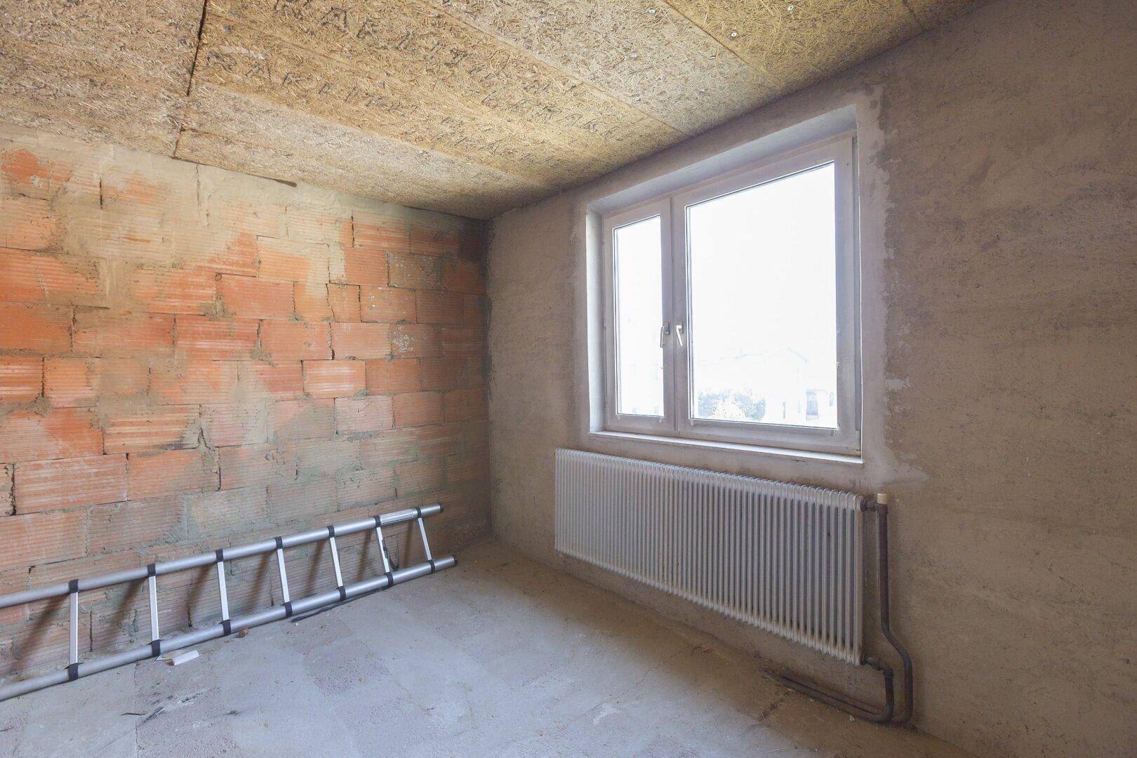 Raum im Rohbau mit unverputzten Ziegelwänden und einem Fenster, das Tageslicht spendet.