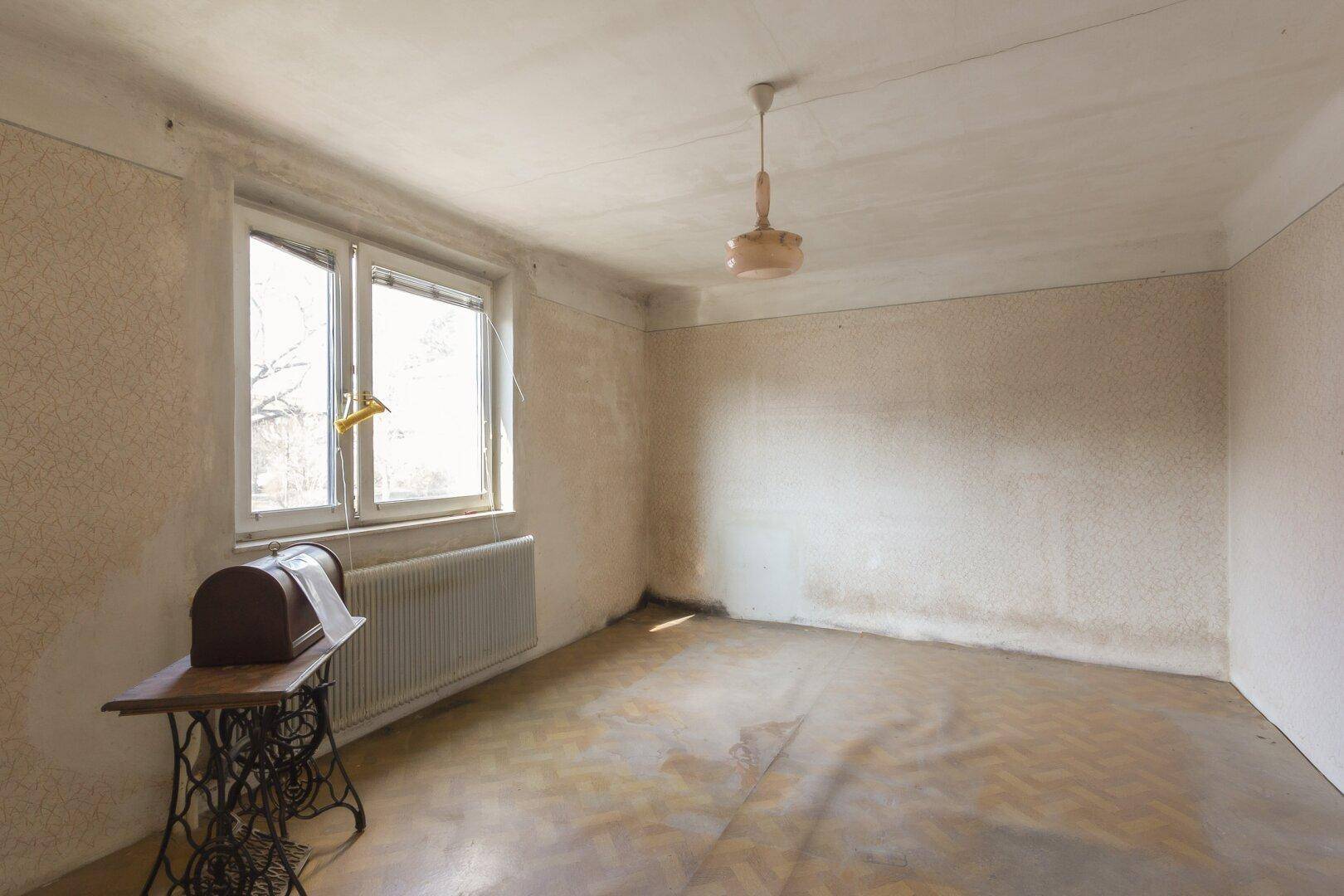 Leeres Zimmer mit abgenutztem Parkettboden und einem Fenster, das Tageslicht hereinlässt.