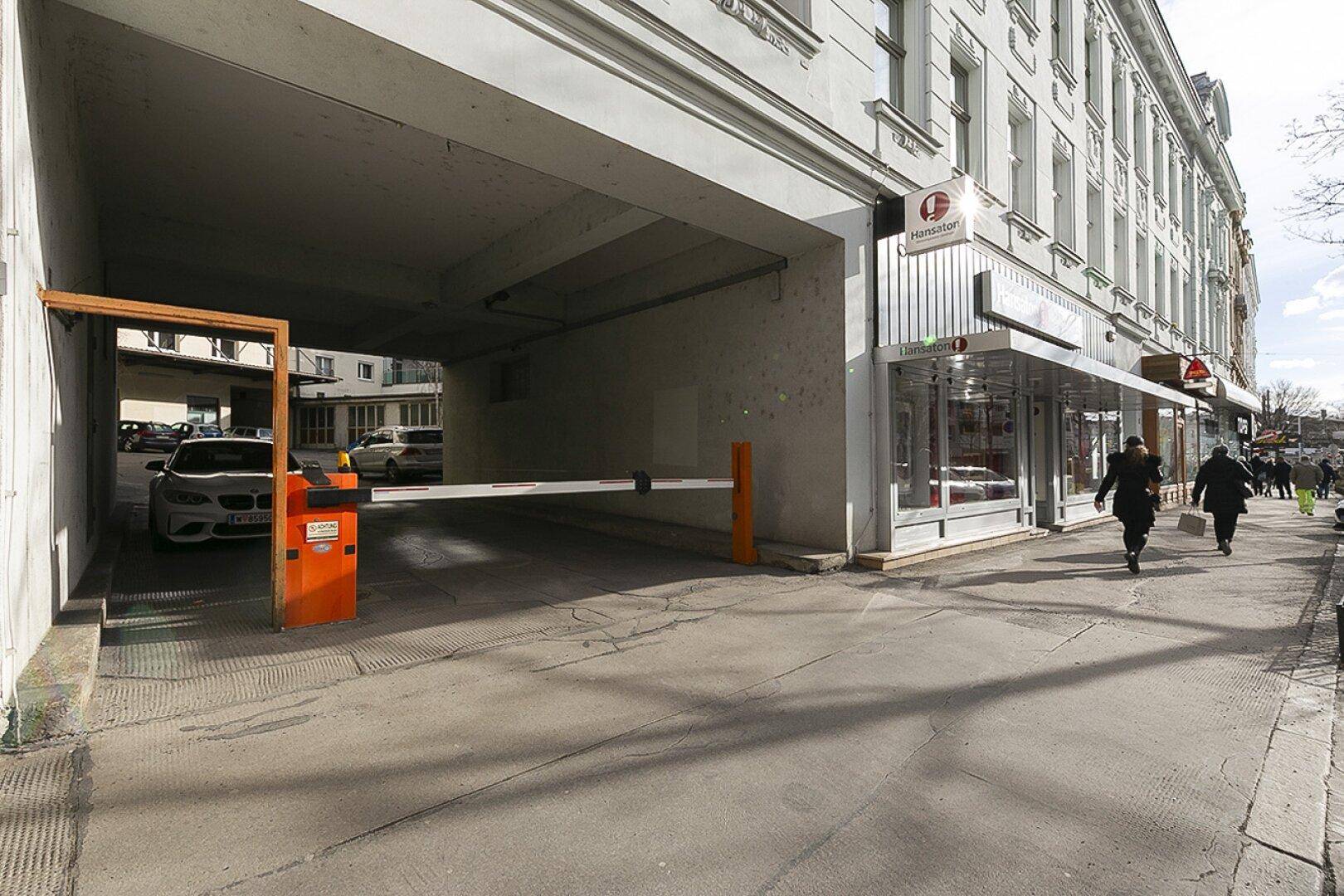Eingang zu einer Tiefgarage oder einem überdachten Parkbereich mit Schranke und angrenzendem Gebäude in städtischer Umgebung.