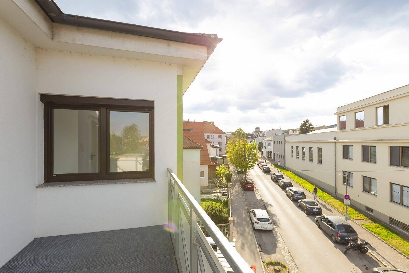 Blick von einem Balkon auf eine belebte Straße mit parkenden Autos und Gebäuden unter einem bewölkten Himmel.