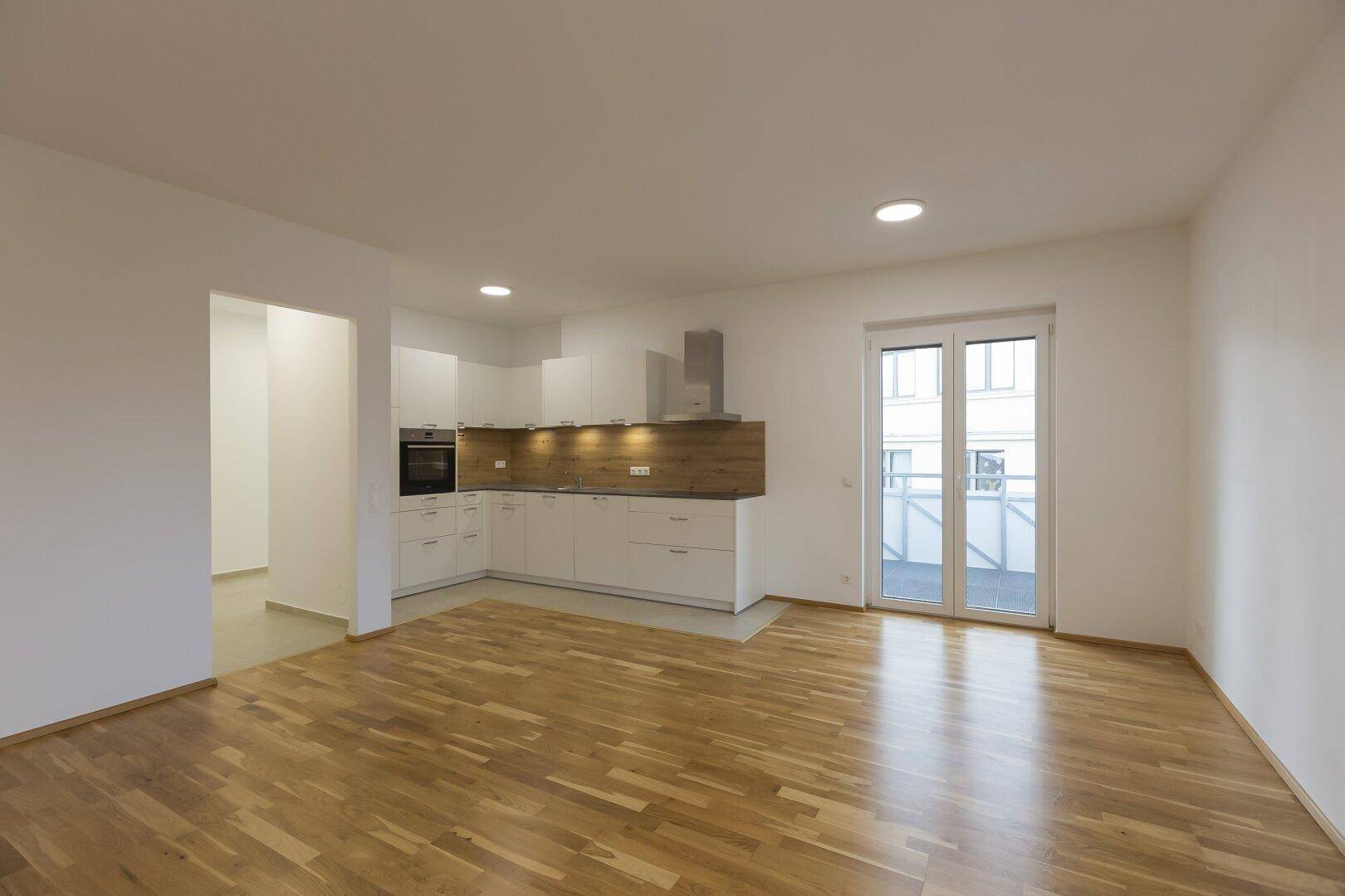 Heller offener Wohnbereich mit neuer Einbauküche, Holzboden und Zugang zu einem Balkon.