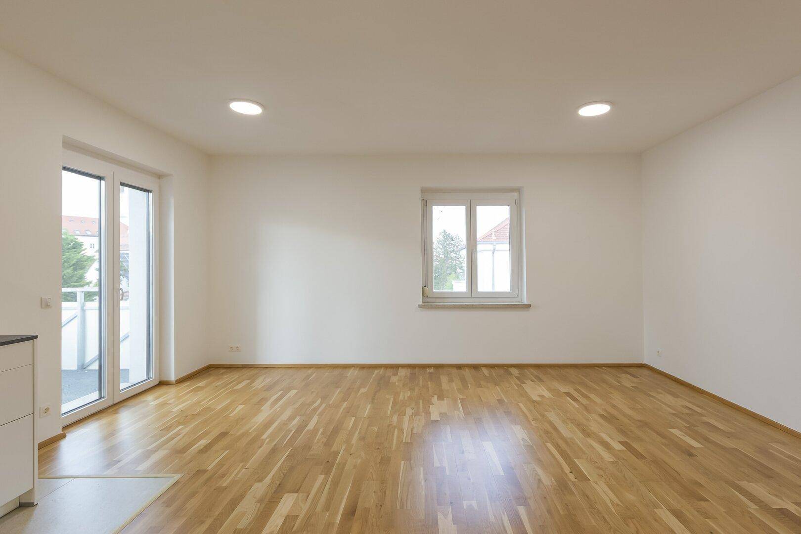 Lichtdurchfluteter Wohnbereich mit hellem Holzboden und großen Fenstern sowie Zugang zum Balkon.