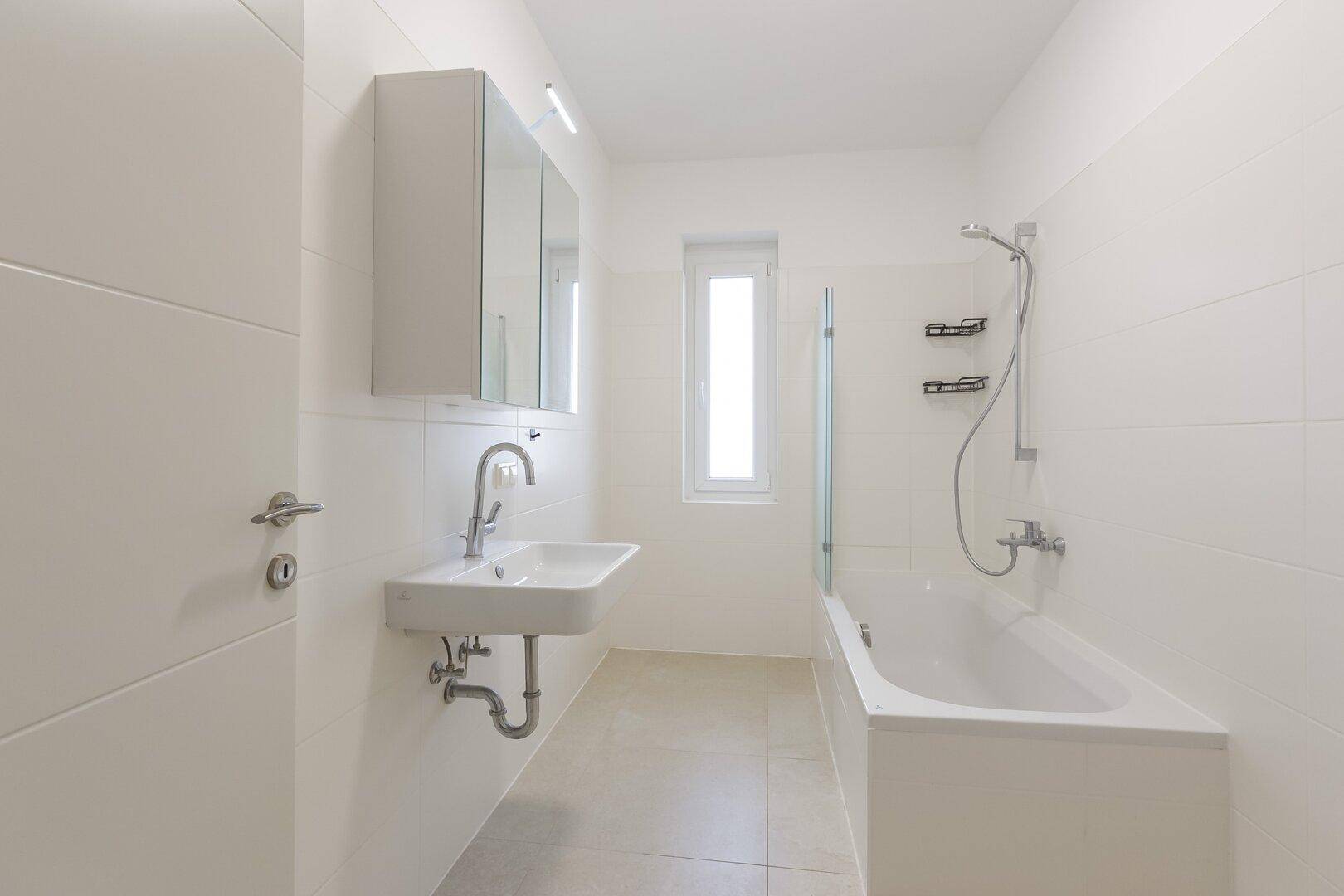 Modernes Badezimmer mit weißen Fliesen, Badewanne mit Duschfunktion, Waschbecken und einem Fenster.