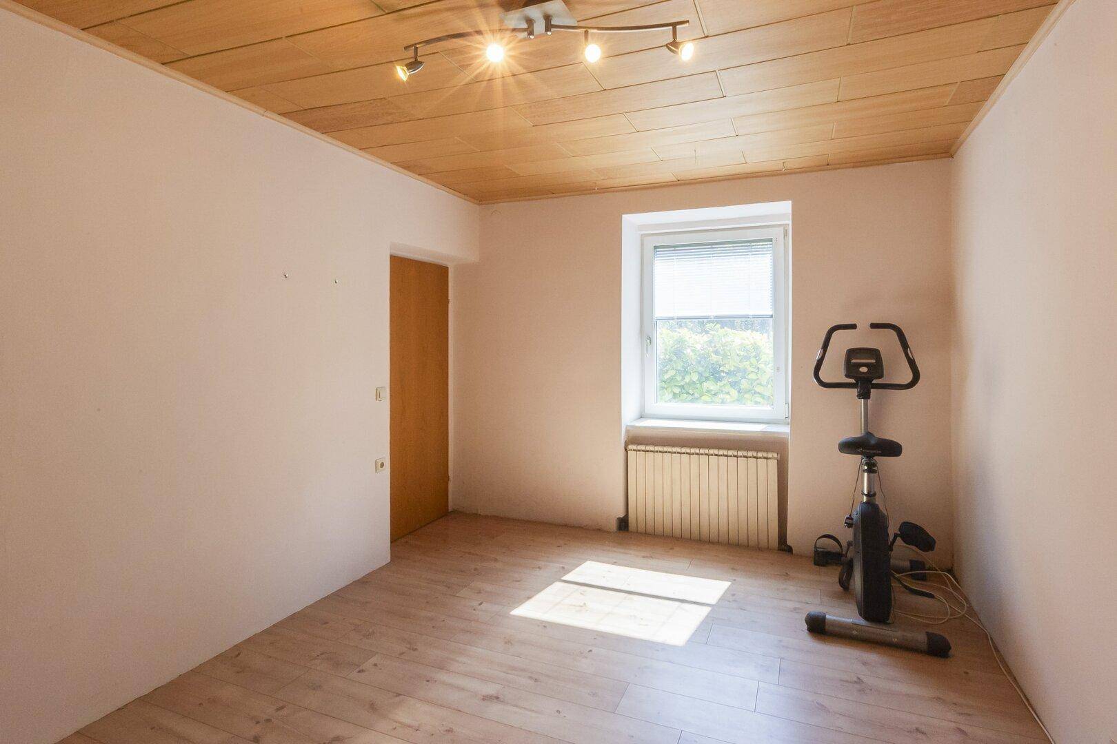 Helles Zimmer mit Holzbalkendecke, Parkettboden und einem Fenster mit Blick ins Grüne.