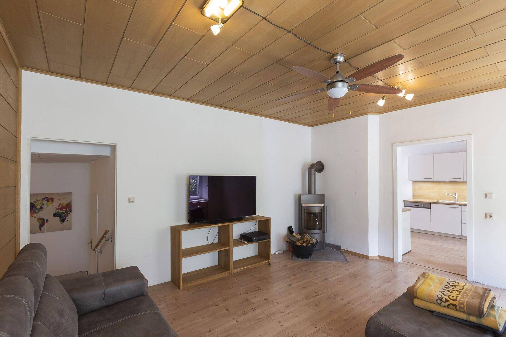 Gemütliches Wohnzimmer mit Holzbalkendecke, Kaminofen und Zugang zur Küche.