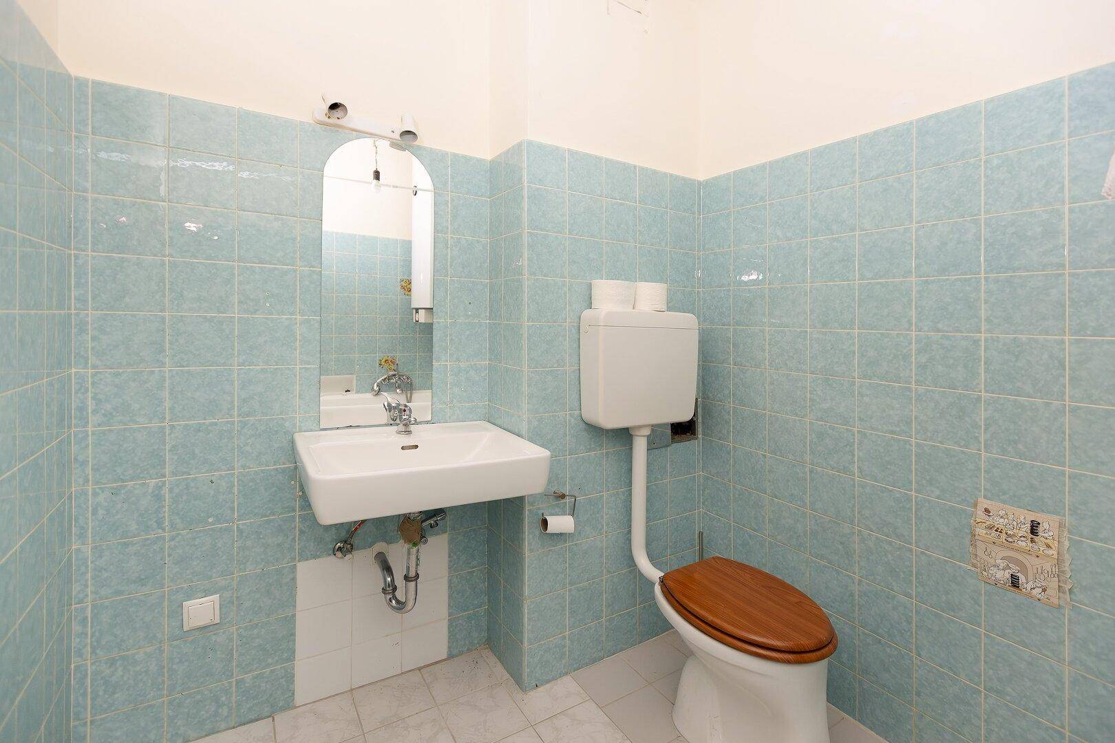 WC mit hellblauen Fliesen, einem Waschbecken und einer Toilette mit Holzdeckel.