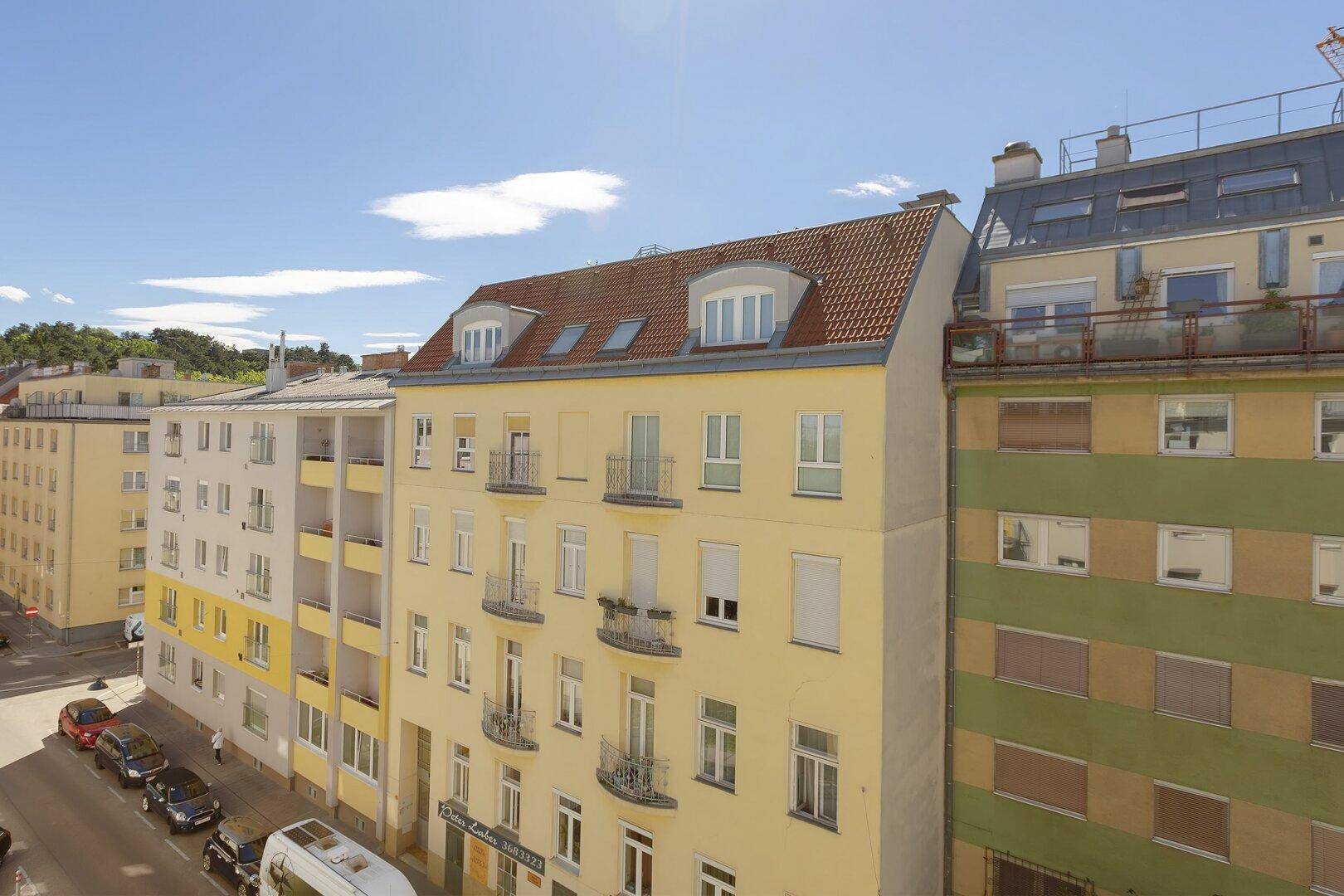 Mehrfamilienhaus mit gelber Fassade, roten Dachziegeln und kleinen Balkonen unter blauem Himmel.