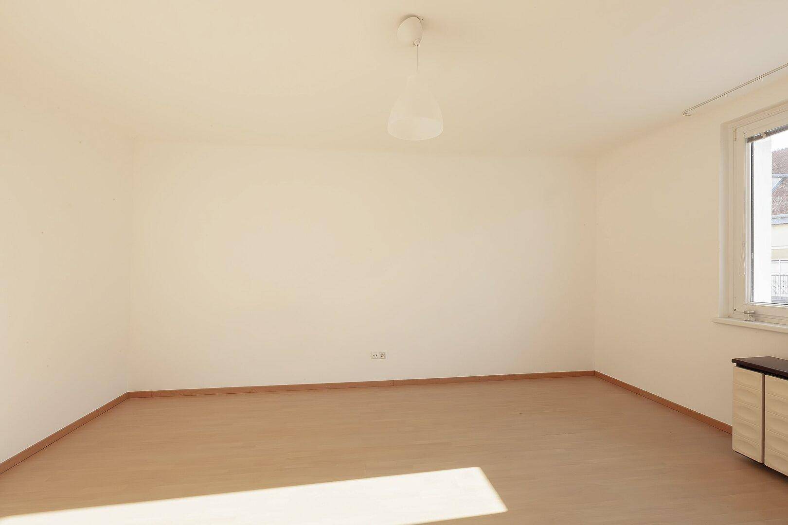 Leeres Zimmer mit hellem Laminatboden und einem Fenster, das den Raum mit Sonnenlicht füllt.