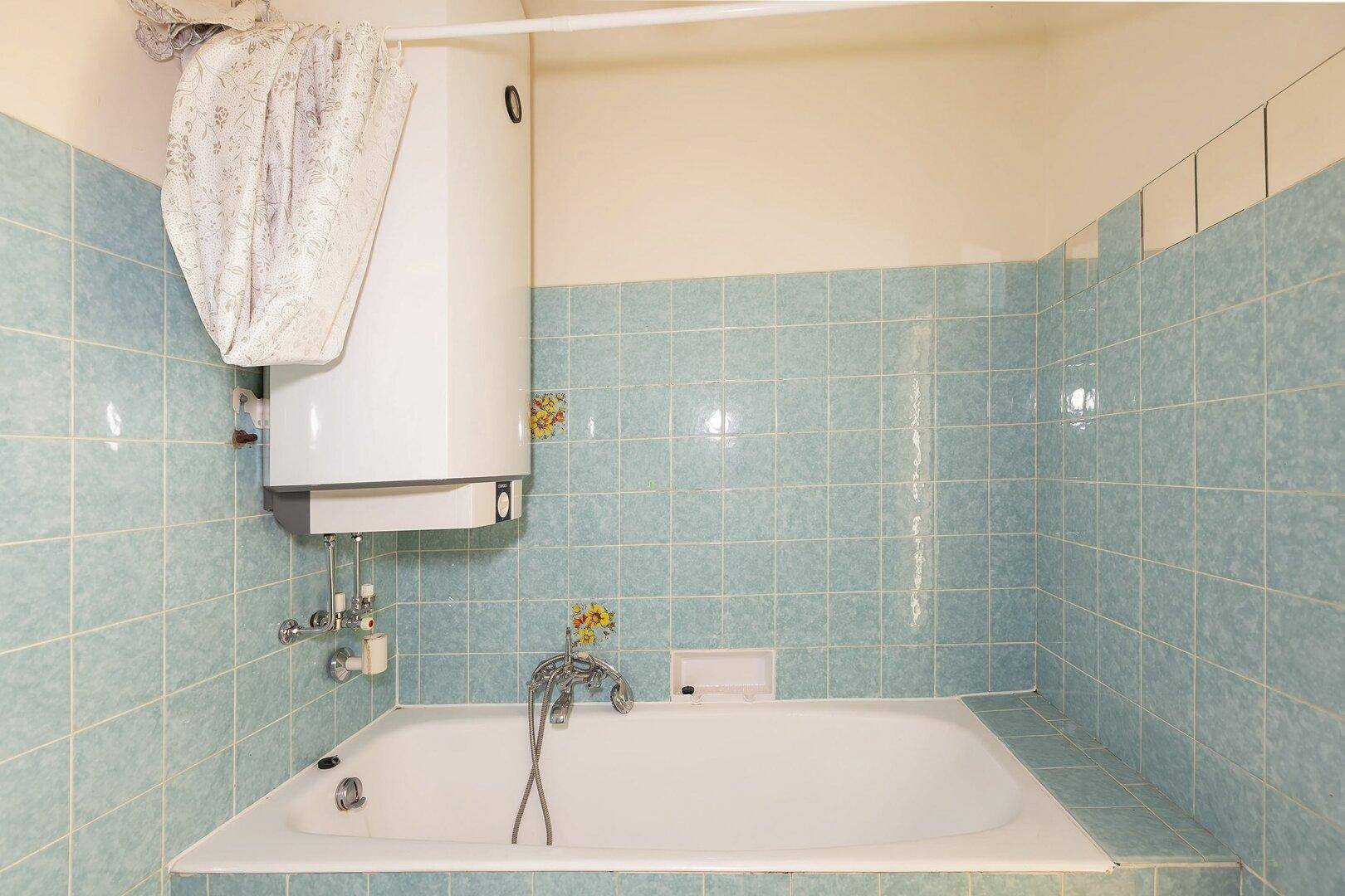 Badezimmer mit hellblauen Fliesen, einer Badewanne und einem Warmwasserboiler an der Wand.