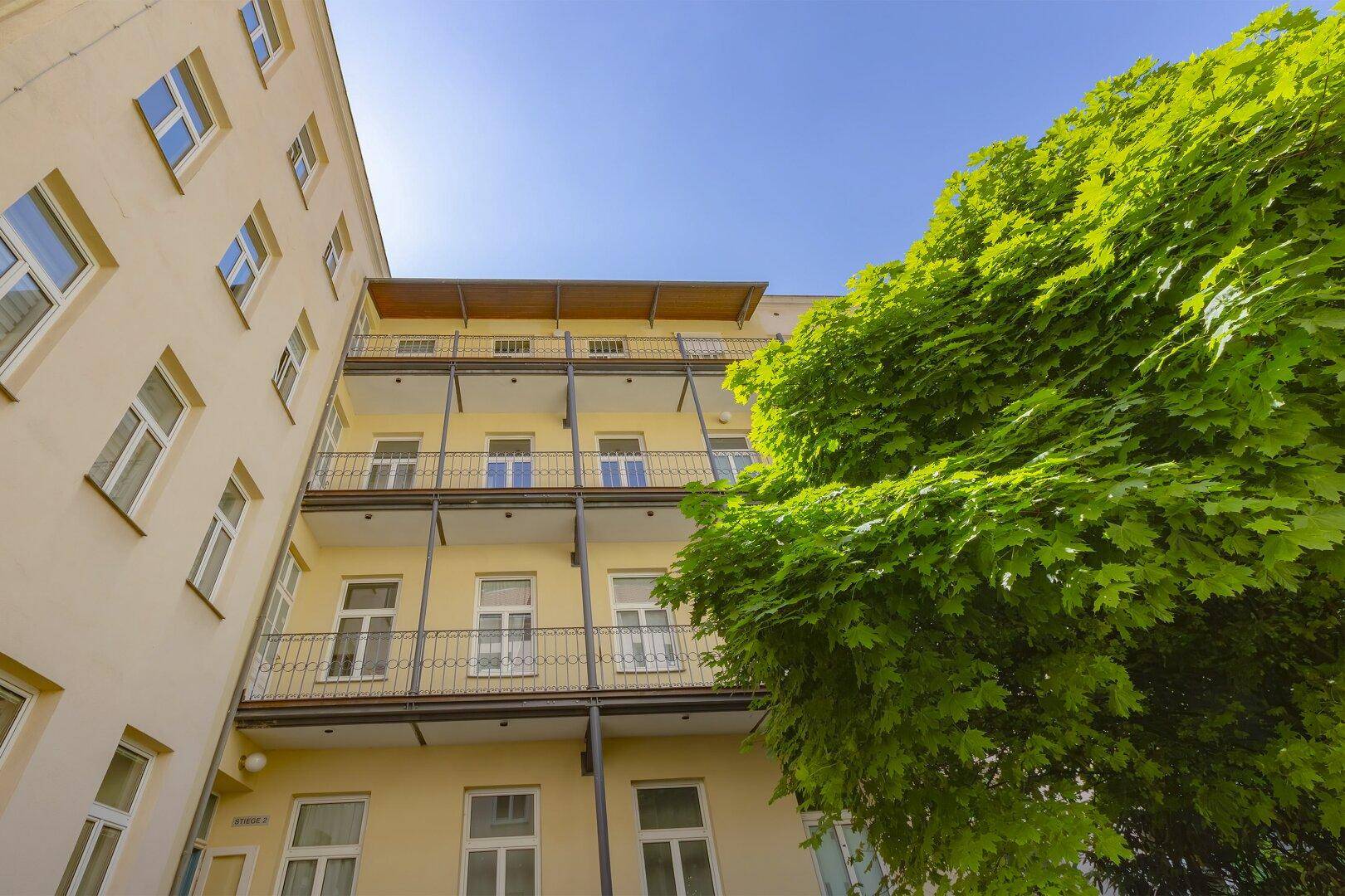 Fassade eines Mehrfamilienhauses mit mehreren Balkonen und einem großen Baum im Innenhof.